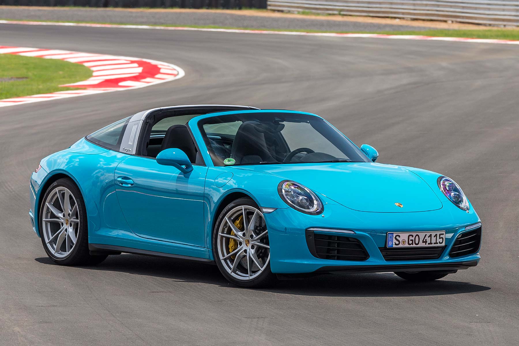 A turquoise Porsche sports car with a person driving is on a racetrack, featuring red and white curbs.