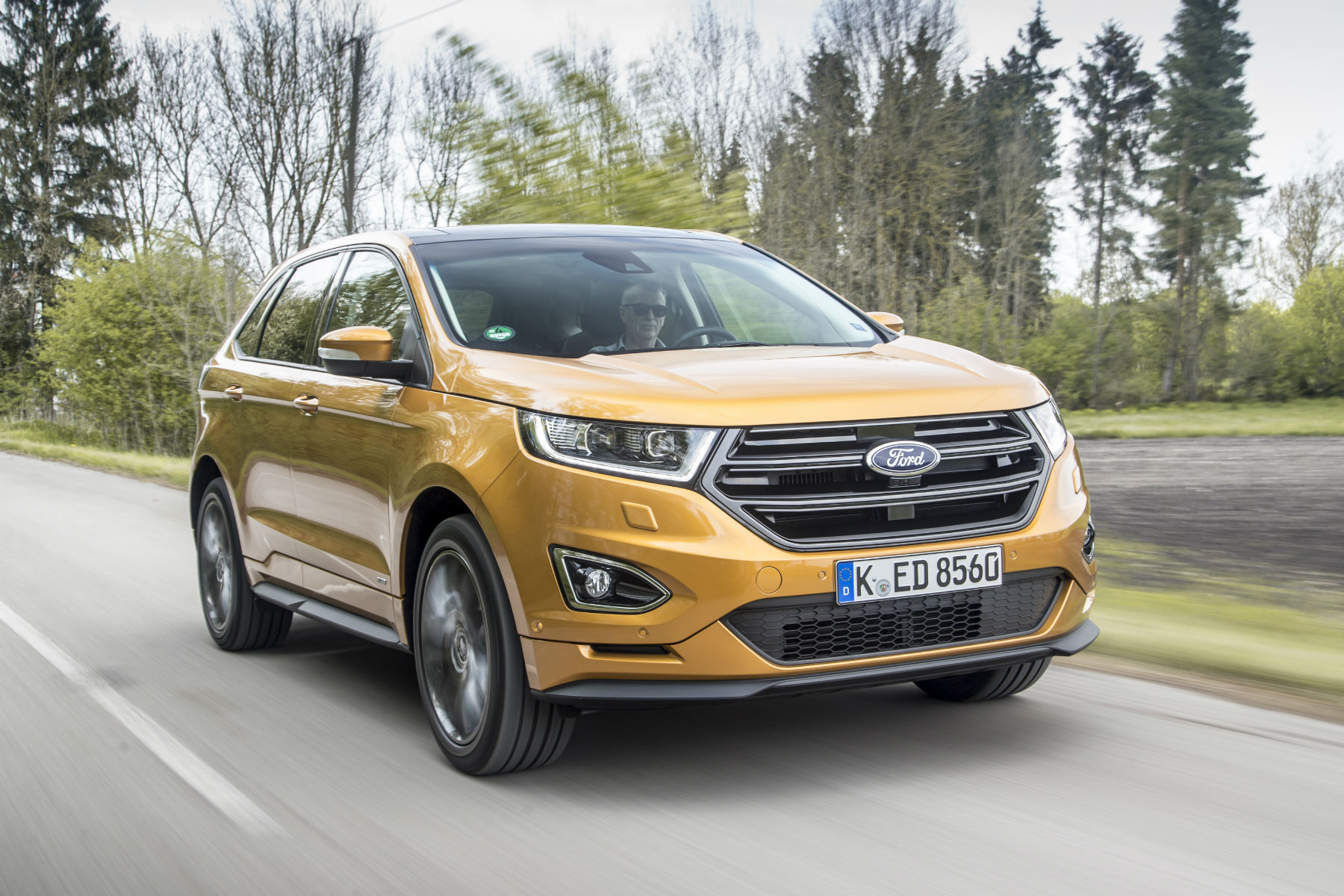 Golden Ford SUV driving on a rural road, surrounded by trees. A person is behind the wheel, wearing sunglasses.