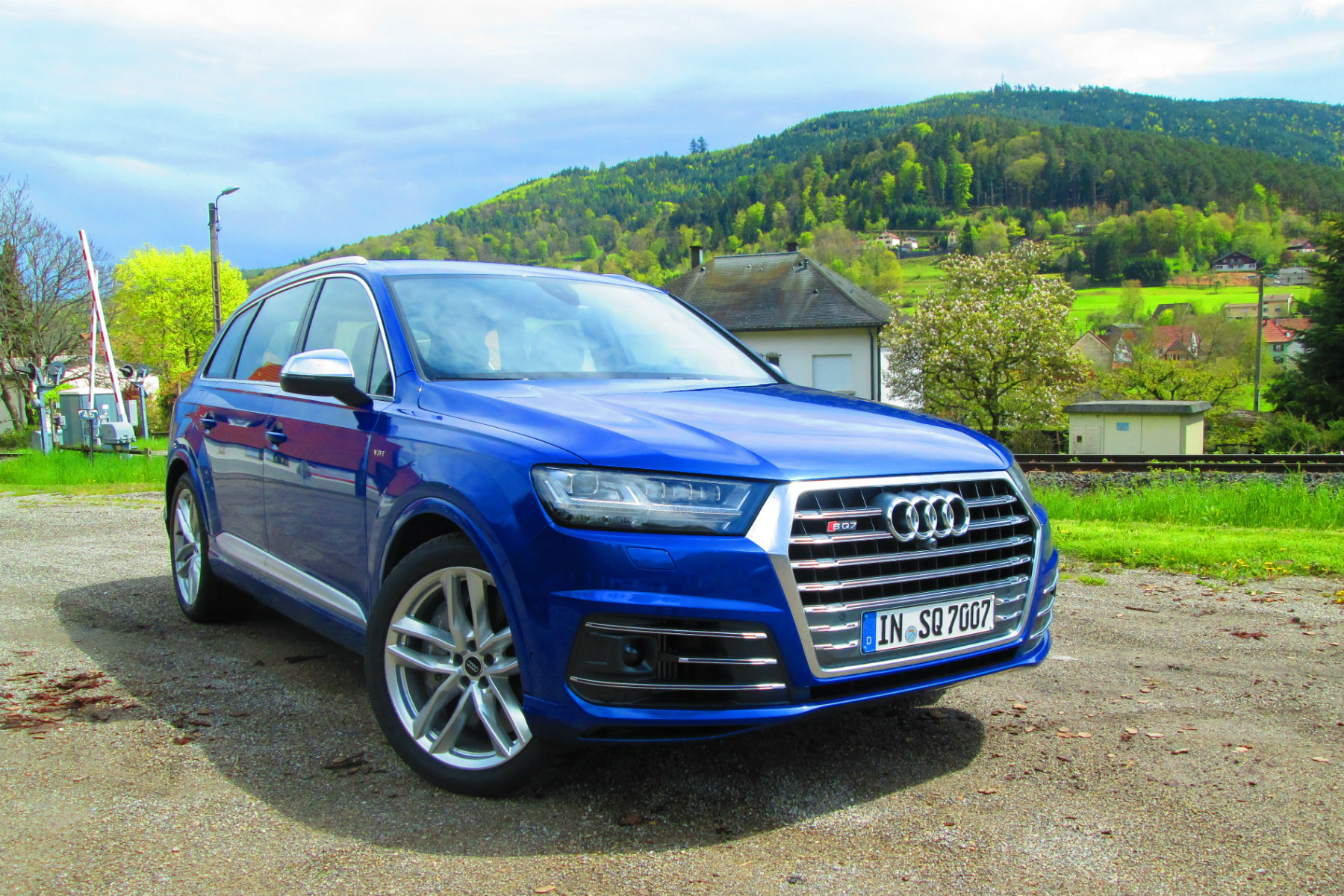 A blue Audi SUV parked outdoors on a scenic rural road, surrounded by lush green hills and scattered buildings in the background.