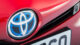 Close-up of a red car's front, highlighting a blue Toyota emblem with partial view of a license plate underneath.