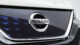 Close-up of a Nissan car front grille featuring a shiny chrome emblem on a dark blue patterned background, showcasing sleek, modern design.