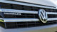 Close-up of a Volkswagen California XXL grill showing emblem and horizontal stripes, reflecting greenery and blue sky in the polished surface.