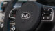 Close-up of a Kia car steering wheel, showing controls and logo, with interior dashboard slightly visible in the background.