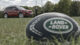 A red Land Rover parked near rugby goalposts with a Land Rover rugby ball in the foreground, surrounded by trees under a cloudy sky.
