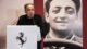 A person speaks at a podium with the Ferrari logo, standing in front of a large, vintage portrait of a smiling person.