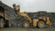 A large yellow loader transfers rocks into a dump truck in a quarry, surrounded by rugged stone cliffs under a cloudy sky.