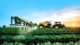 A tractor sprays crops in a lush green field at sunrise, with a clear blue sky and distant horizon.