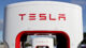 Close-up of Tesla Supercharger station against a blue sky. The brand name is prominently displayed in red on a white background.