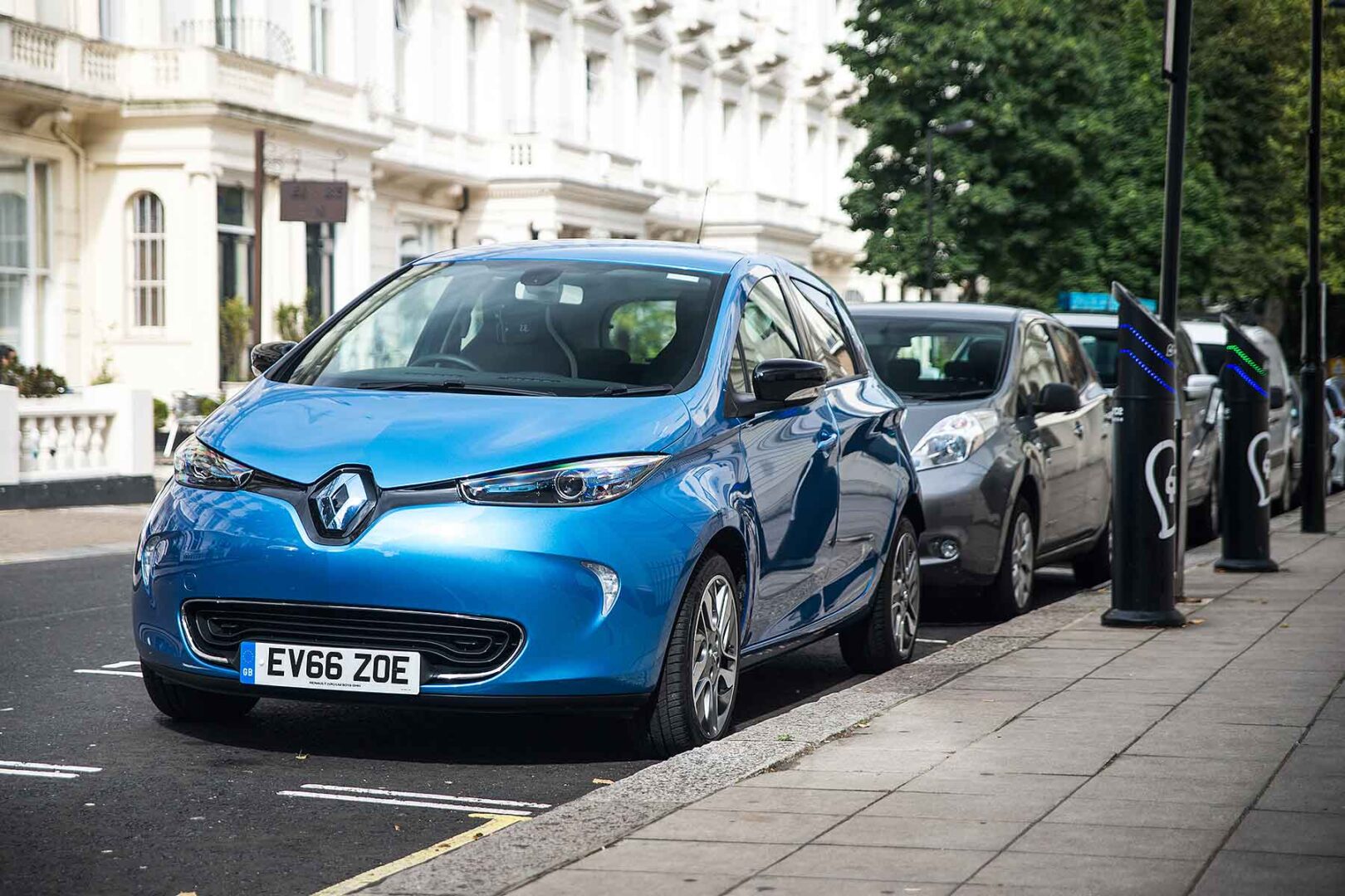 A blue Renault car parked on a street with elegant white buildings, next to electric charging stations and leafy trees.