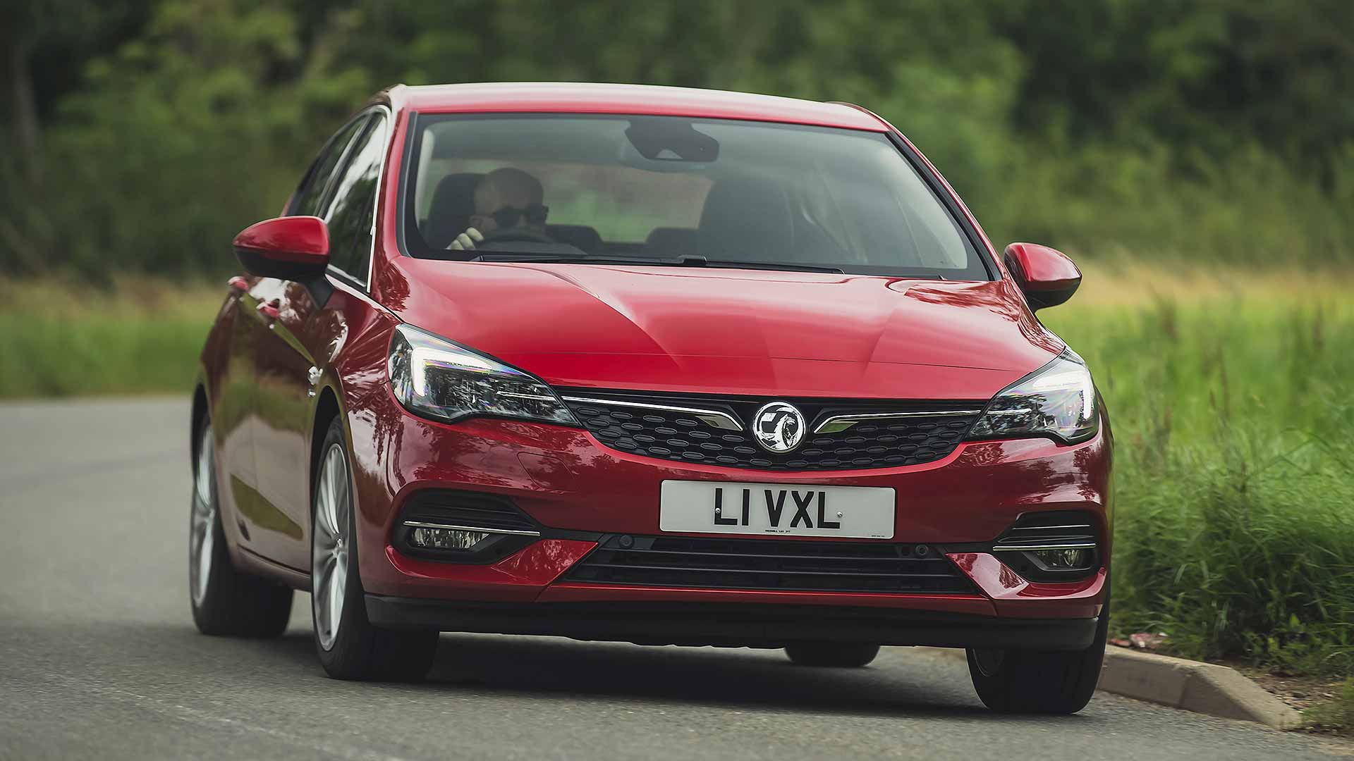 A red Vauxhall car drives on a curving road surrounded by greenery. A person is visible inside, blurred background.