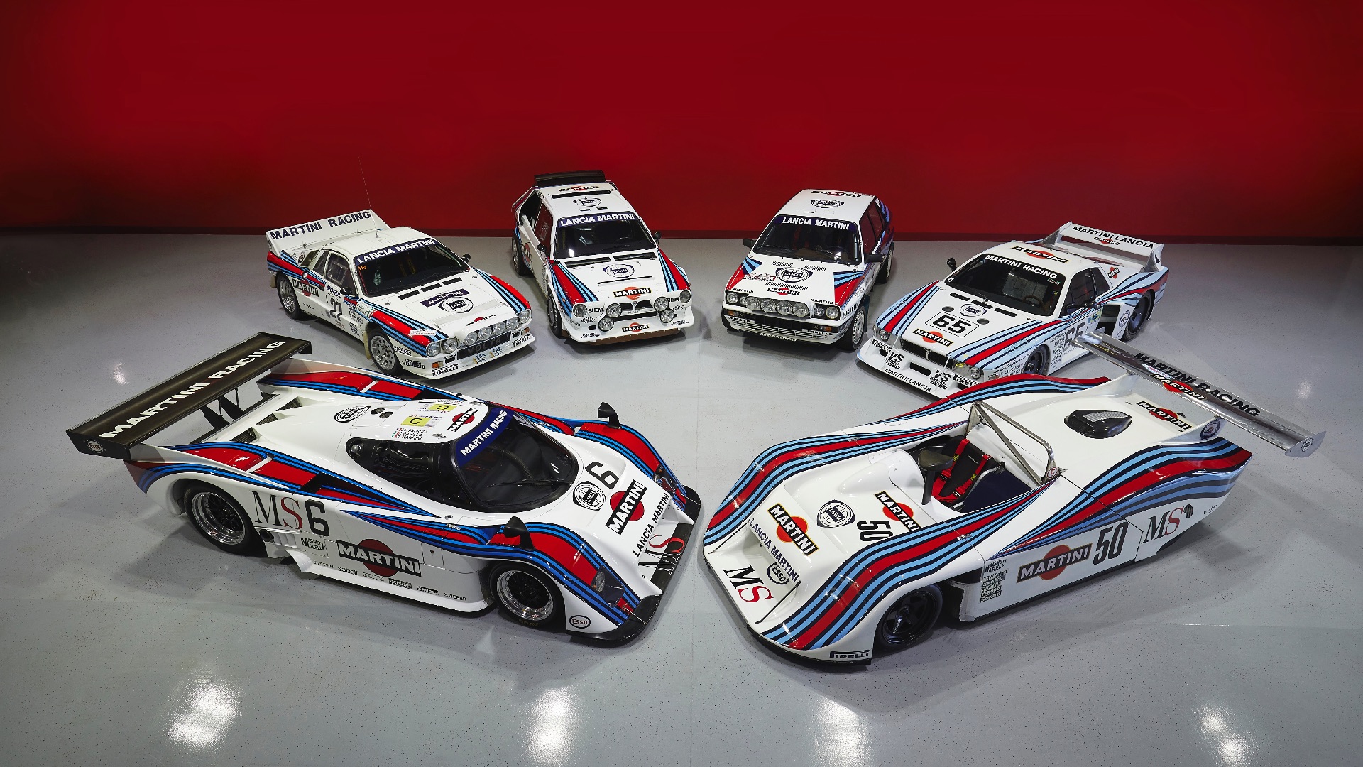 Six classic Martini Racing Lancia cars displayed on a showroom floor with a bold red background, highlighting their iconic livery.