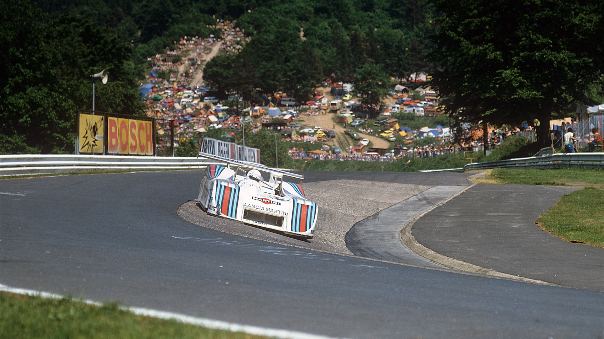 Race car on curved track surrounded by trees and spectators, with tents in the background. Bosch advertisement visible on fence.