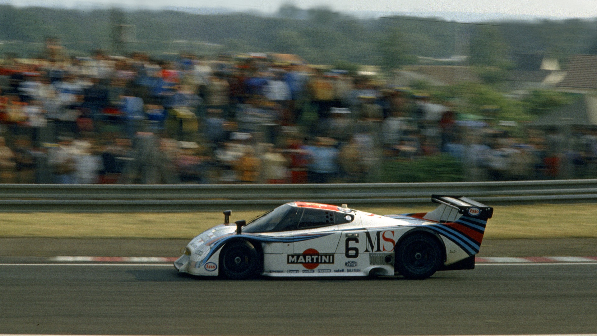 A race car speeds past a crowd of blurred spectators at a racetrack, showing dynamic motion and excitement on a sunny day.