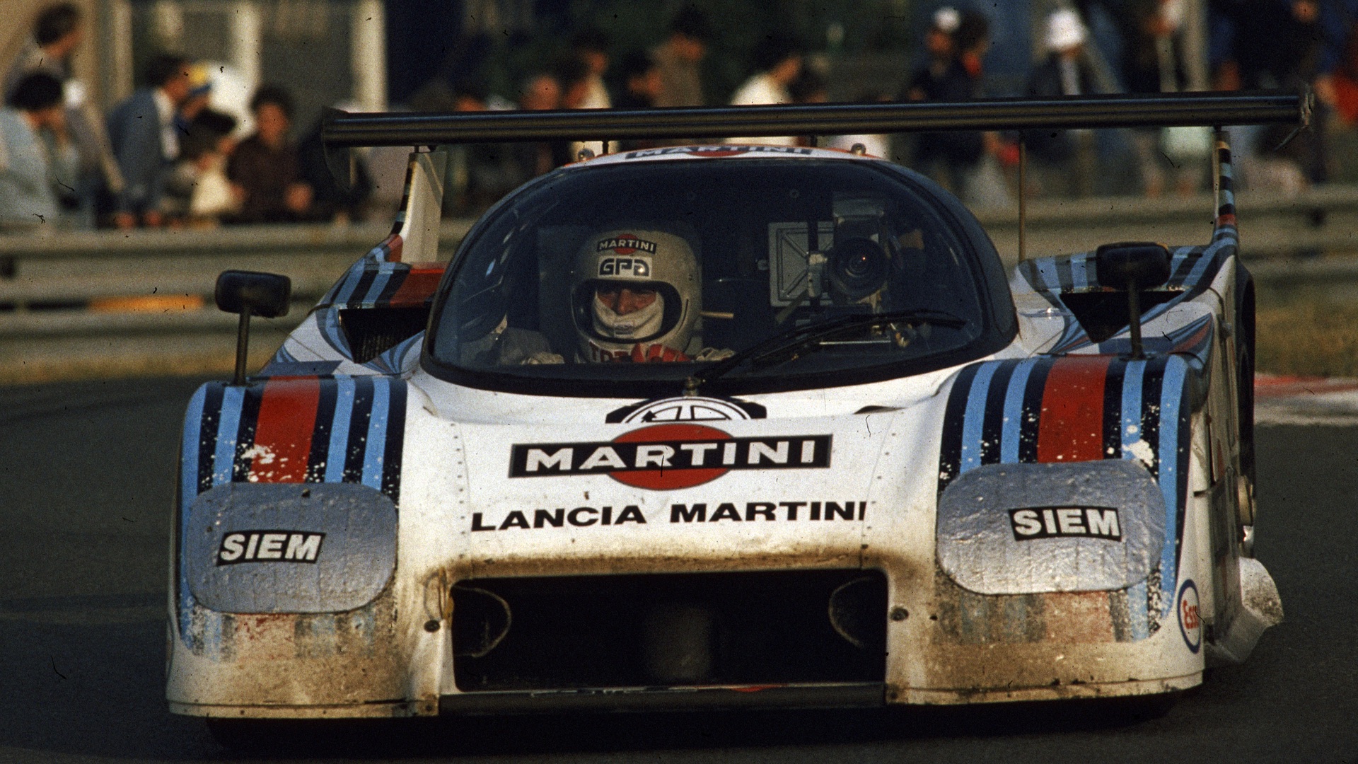 A racing car with Martini branding speeds along track, driver inside. Crowd watches from behind barriers, capturing intense motorsport action.