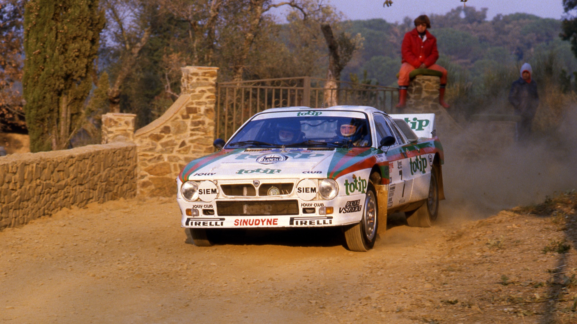 A rally car speeds on a dirt road by a stone wall. Two people watch, one sitting and one standing.