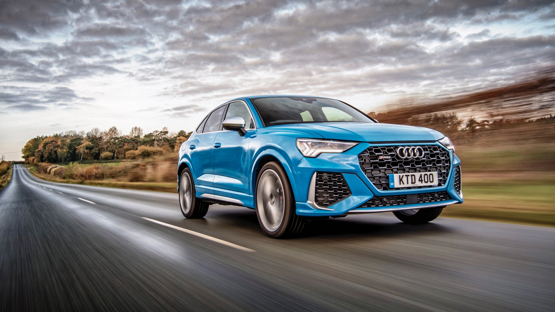A blue Audi car speeds down a rural road, surrounded by fields and trees under a cloudy sky. The scene is dynamic and energetic.