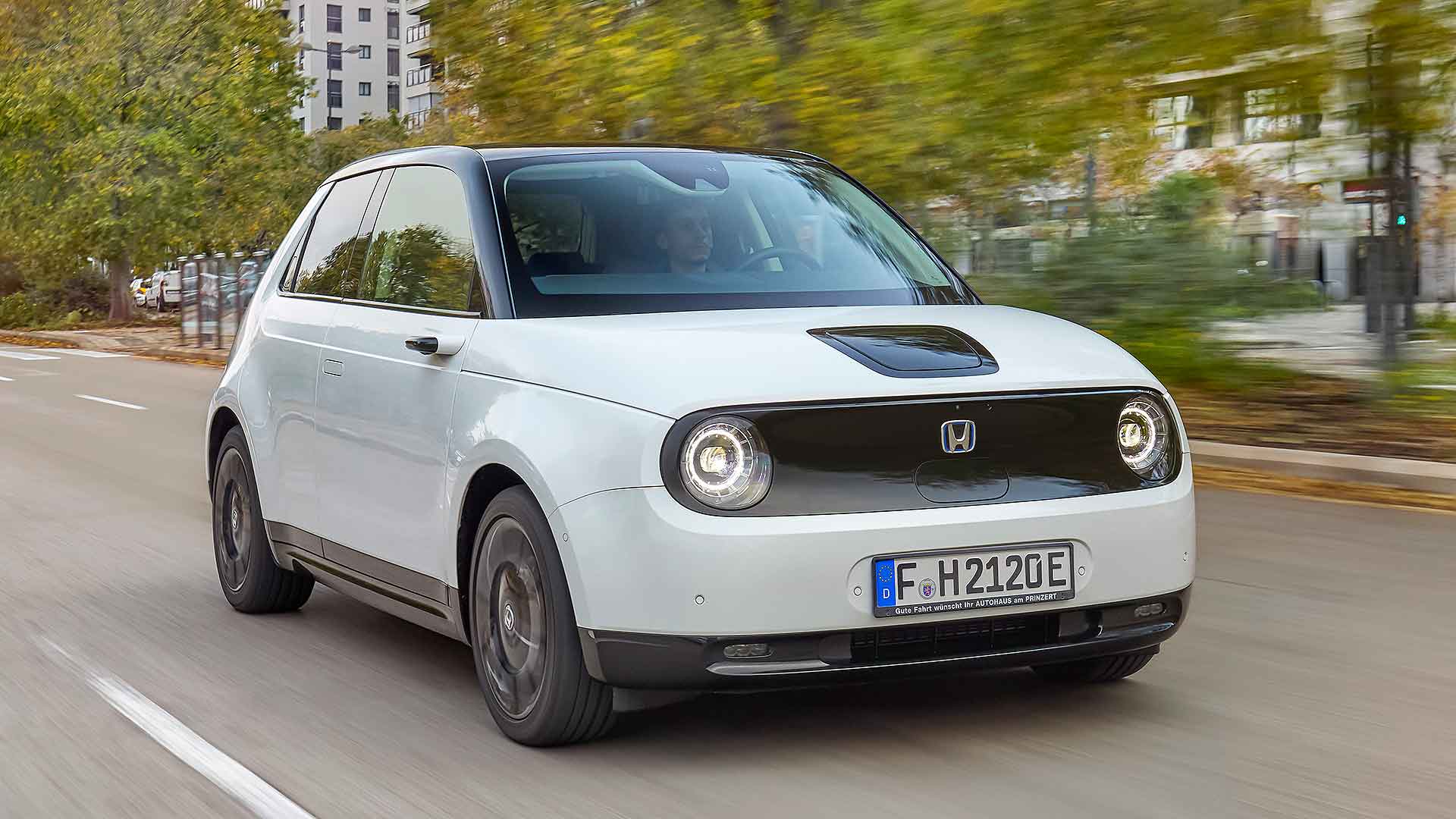 7223_Hondae-17 A white Honda car drives on a city street, surrounded by trees and buildings, with a person visible inside.