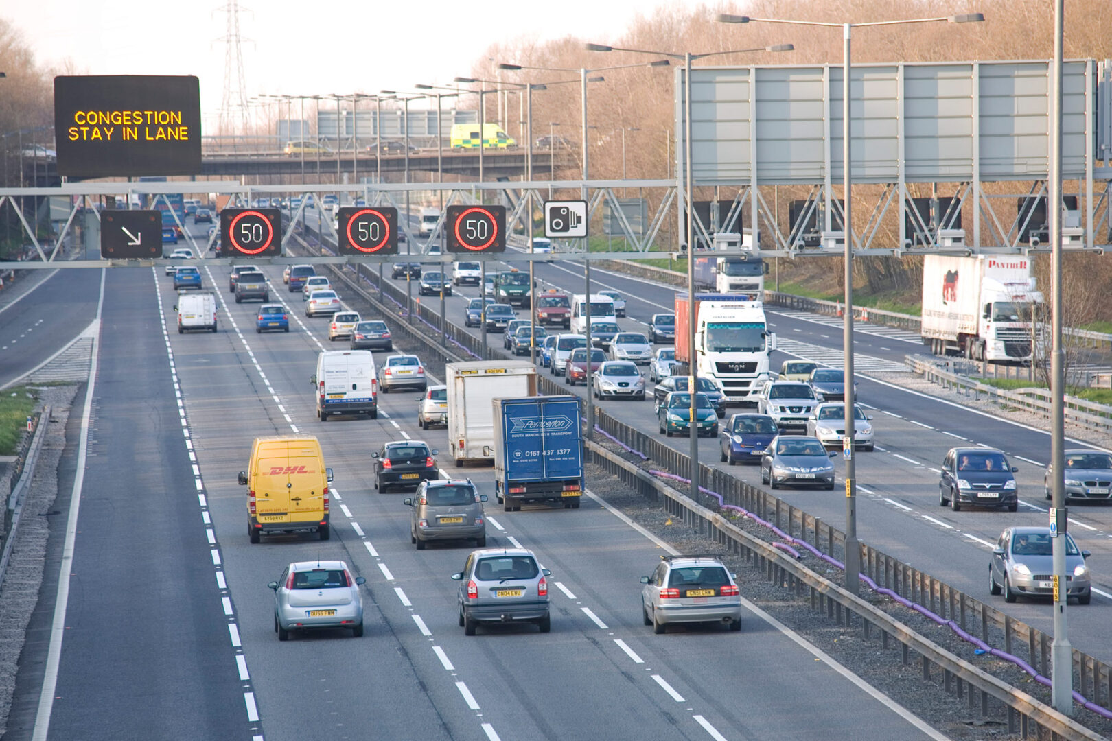 A busy highway with heavy traffic, multiple vehicles, and a congestion warning sign. Overhead gantries display speed limits. Trees line the background.