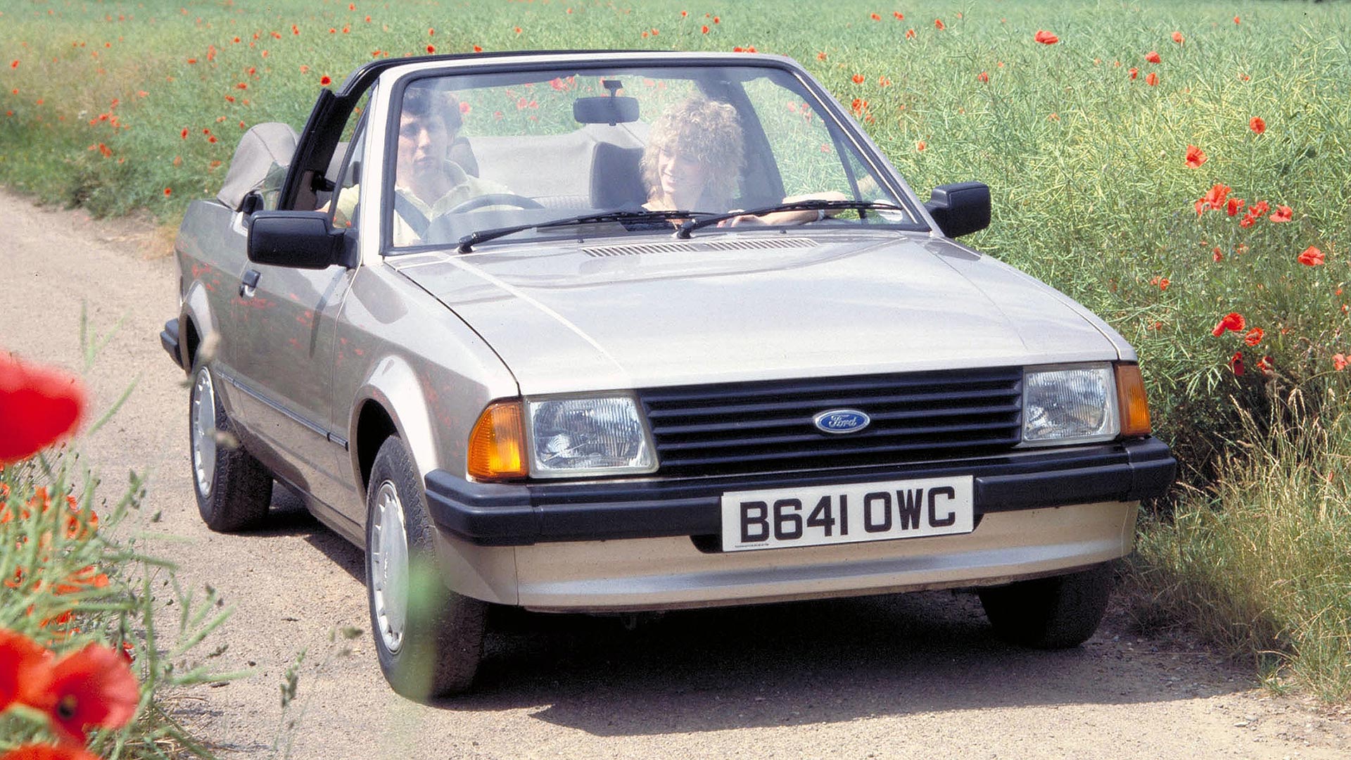 A convertible car on a rural road, surrounded by poppies. Two people inside, enjoying a peaceful sunny day in the countryside.