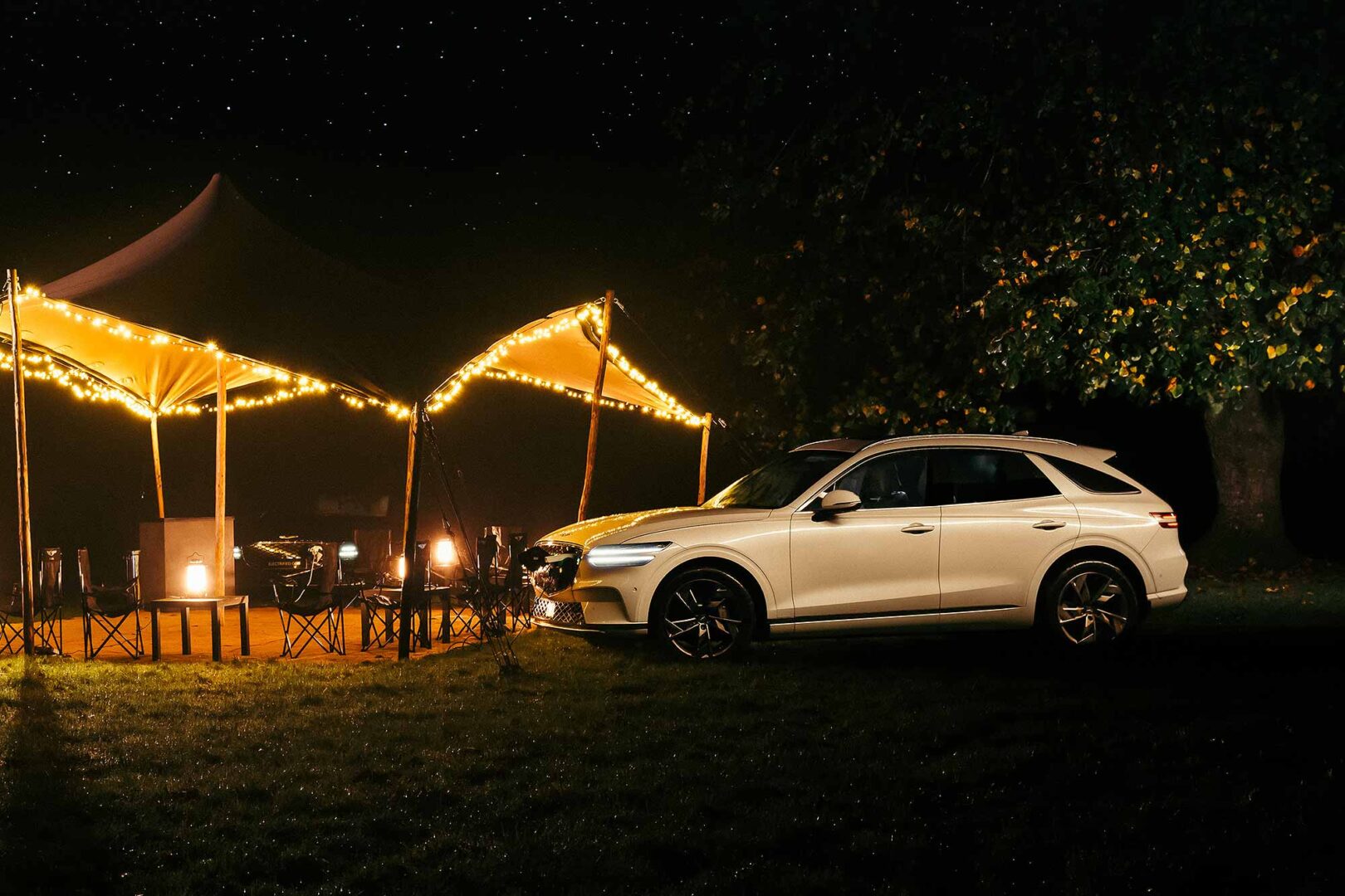 A car is parked next to a lit tent under a starlit sky, surrounded by trees and grassy ground.
