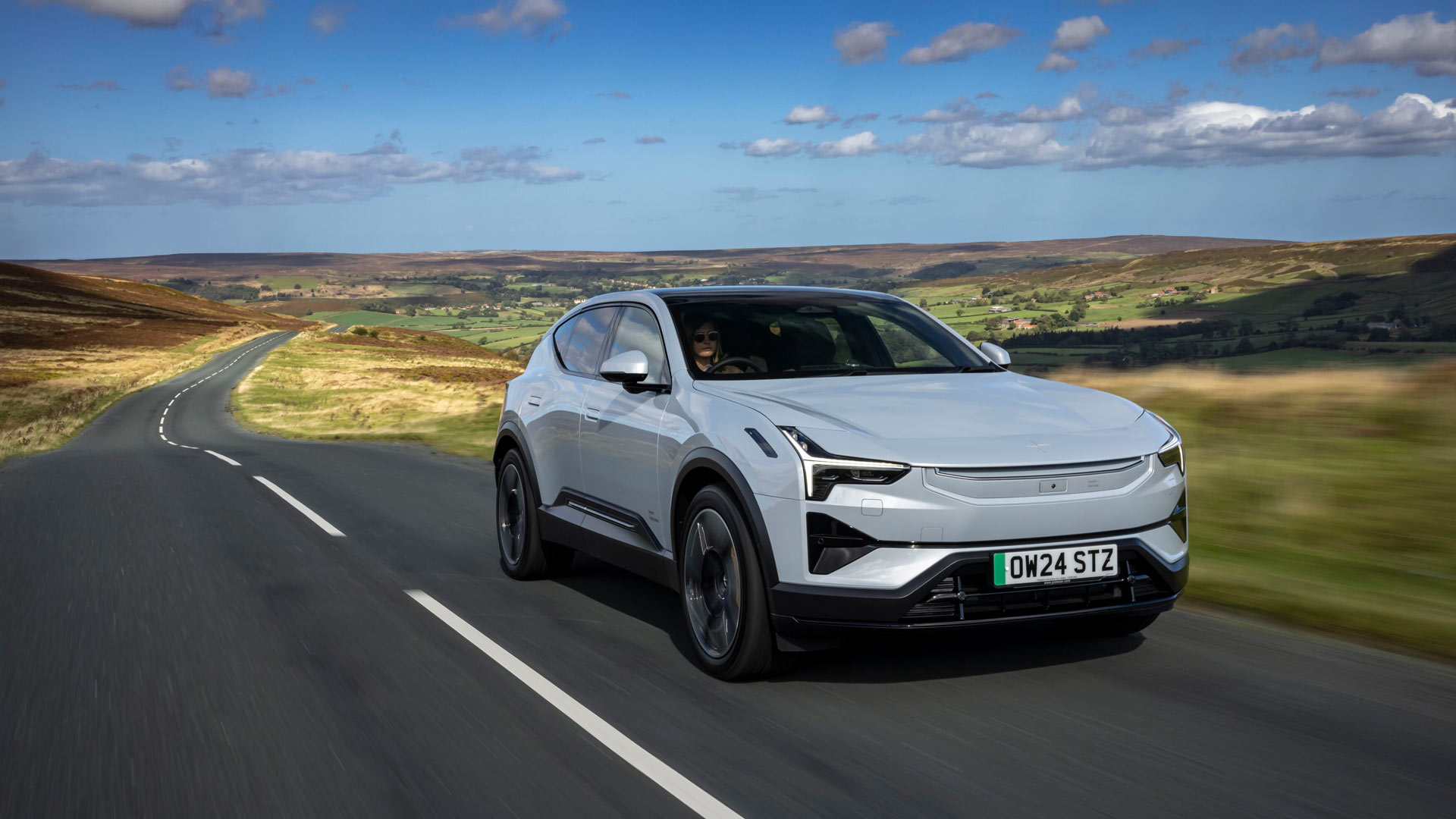 A Polestar 3 car driven by a person on a scenic open road, surrounded by rolling hills and clear skies.