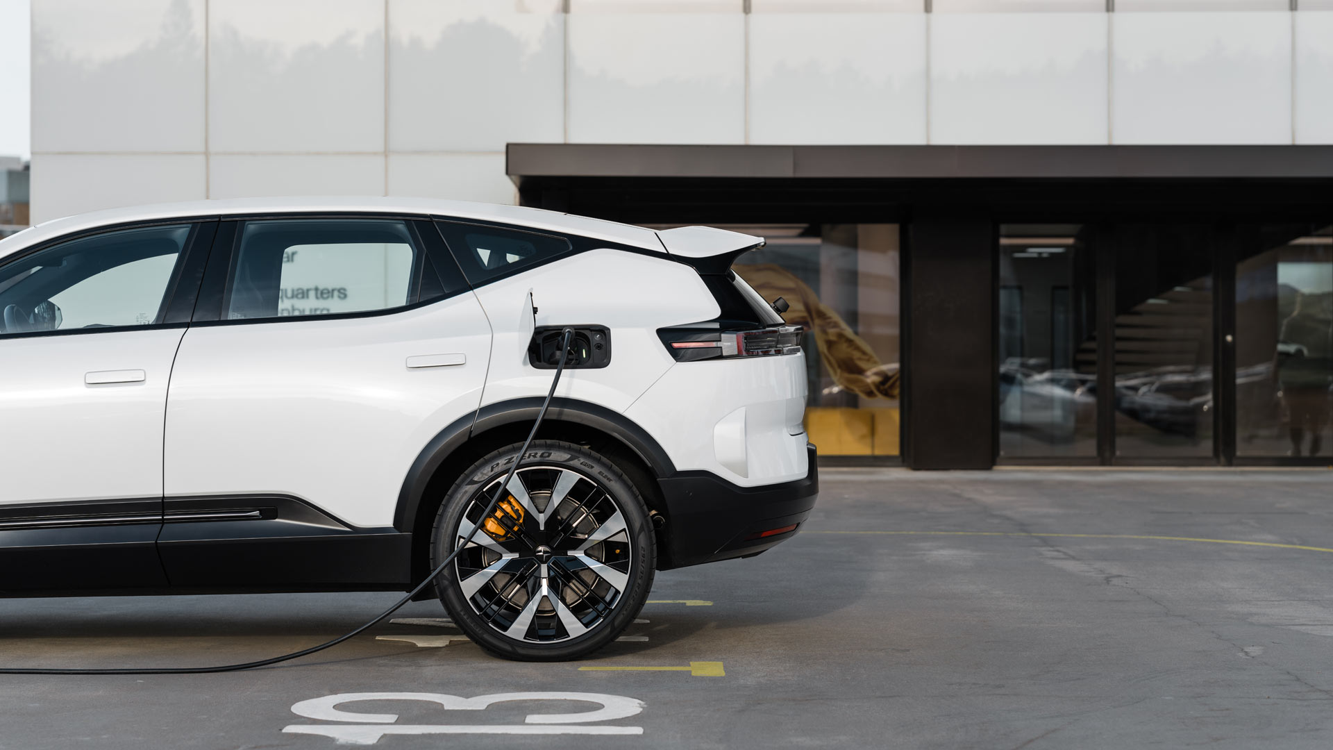 A white Polestar 3 electric car is charging in a parking lot outside a modern building with glass windows and a dark entrance.