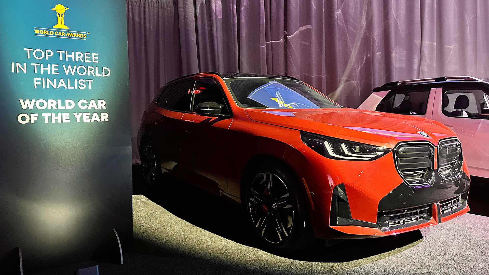 A red BMW vehicle displayed alongside a "World Car Awards" sign, indicating it's a finalist for World Car of the Year.