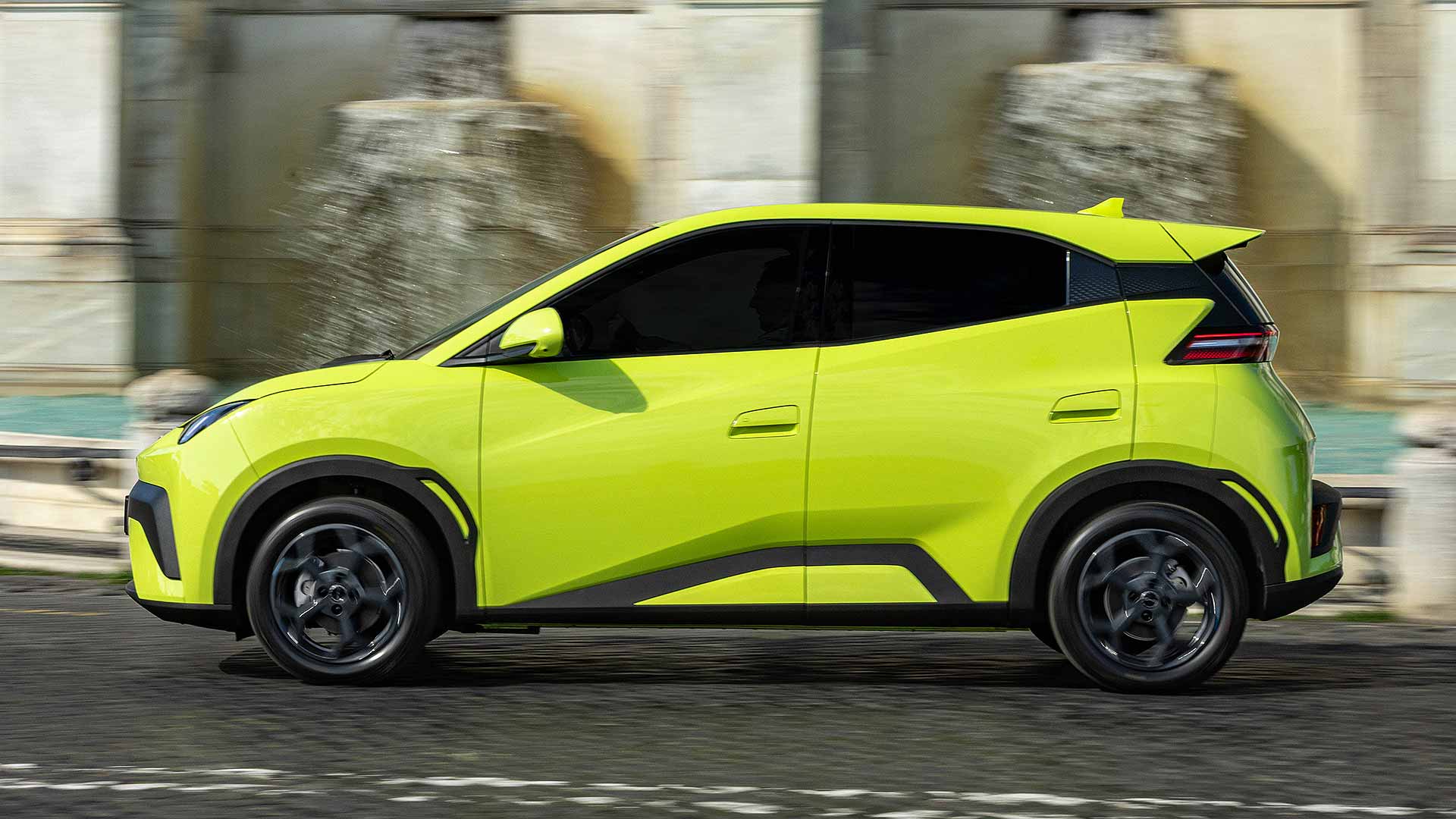Bright green car driving past a stone fountain. Blurred background creates sense of motion and urban setting. No recognizable landmarks visible.