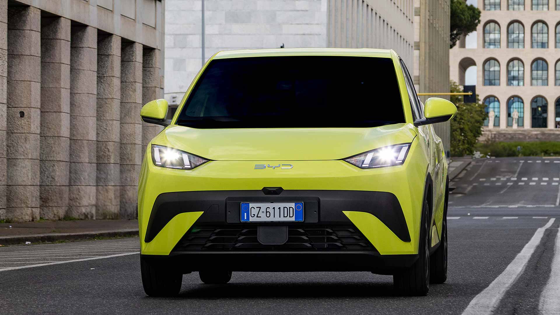 A bright yellow car drives down a city street near Rome's Palazzo della Civiltà Italiana, a building with arched windows.