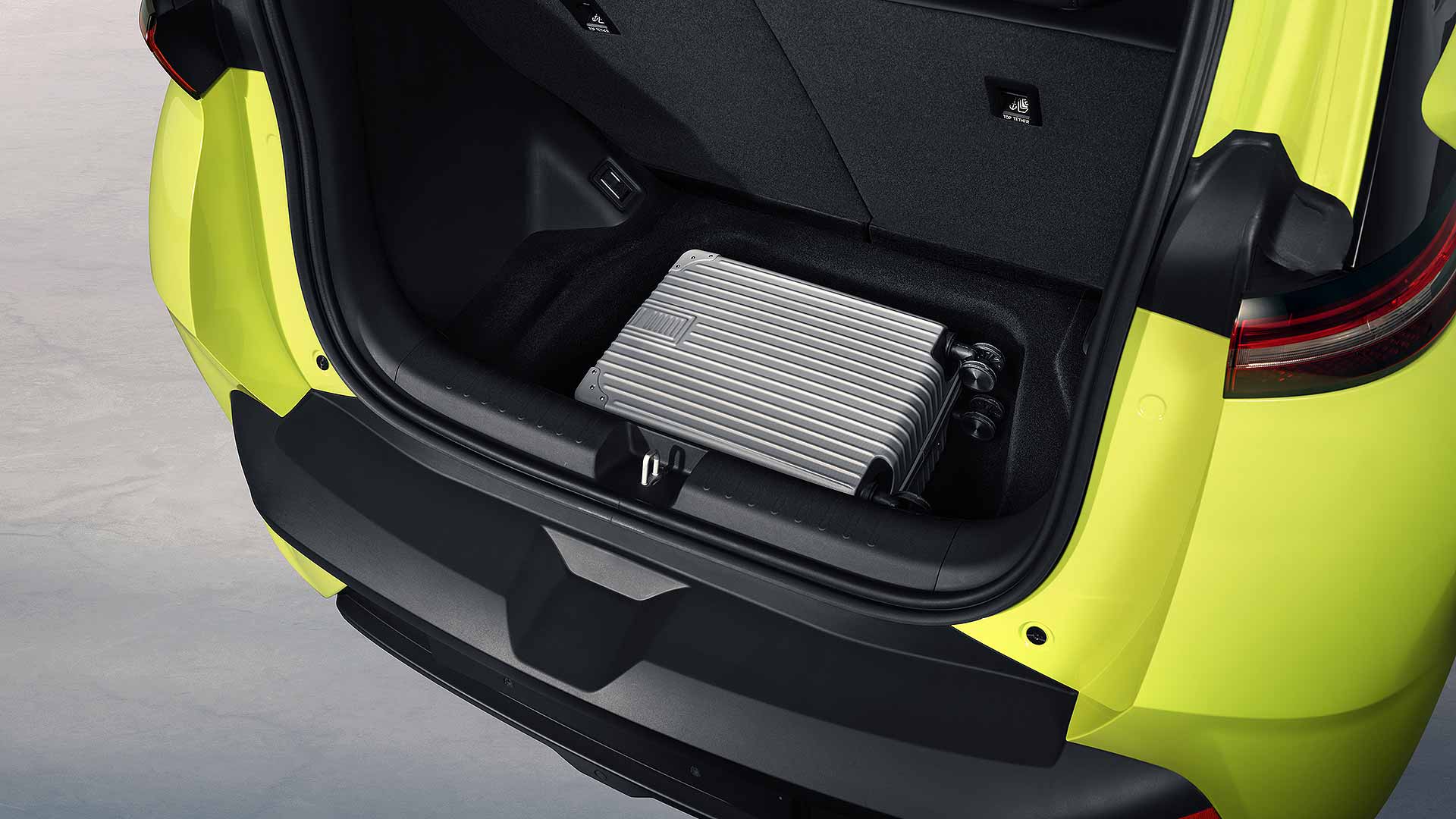 A bright yellow car trunk contains a metallic suitcase. The black interior contrasts with the vibrant exterior, highlighting the vehicle's design.