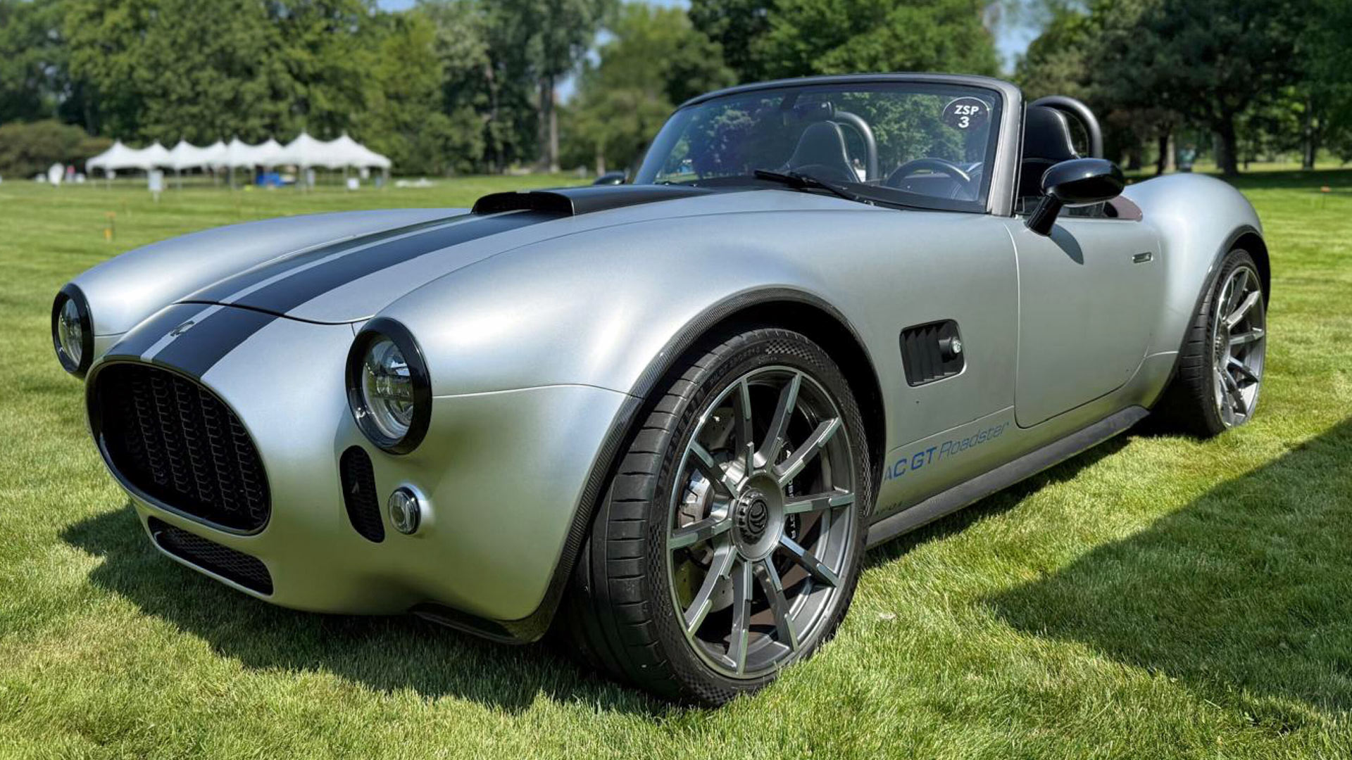 A sleek silver AC GT Roadster parked on lush grass under clear skies, with white tents in the background suggesting an event.