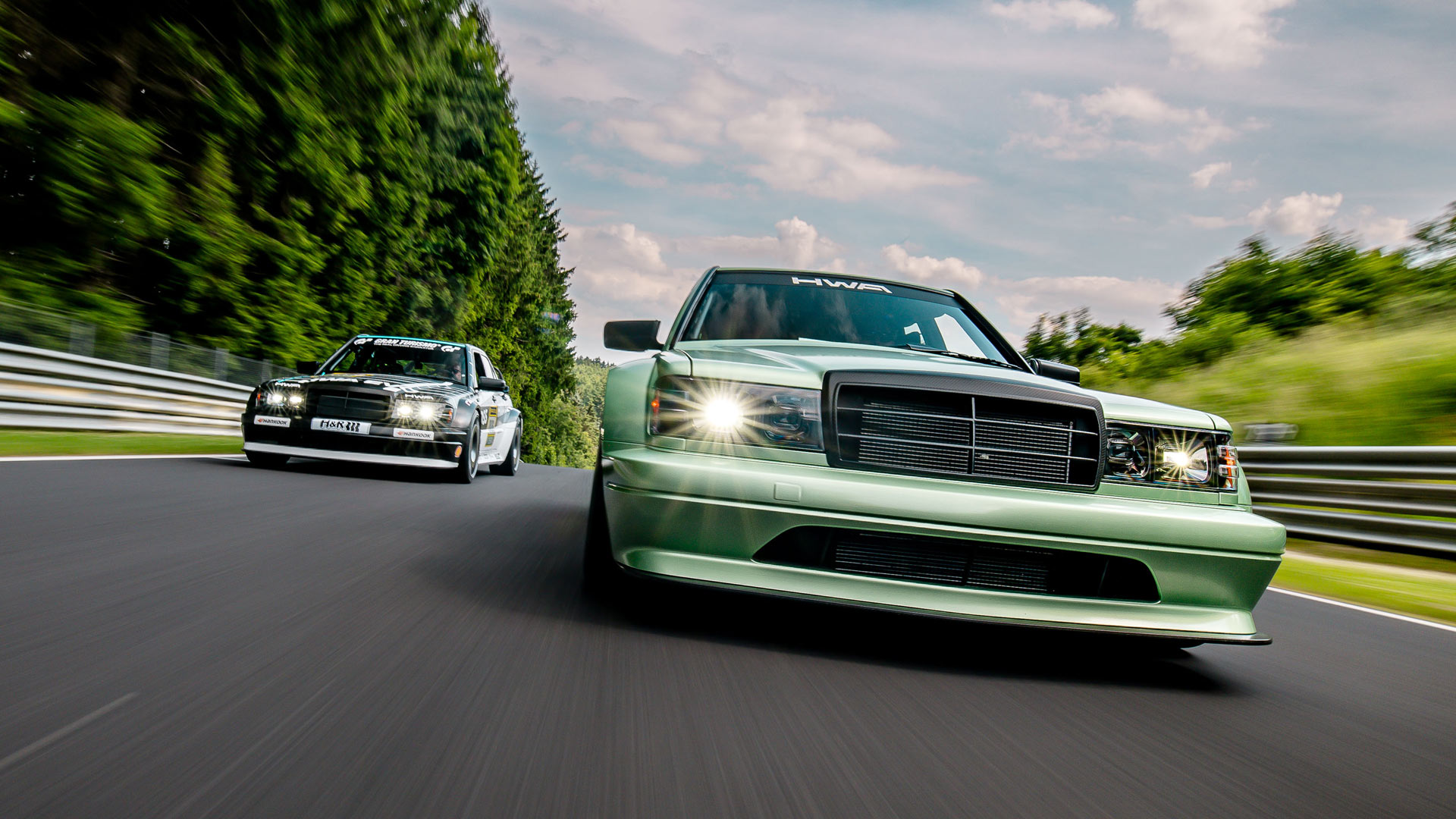 Two HWA Evo cars race on a forested track, headlights on, surrounded by green trees and a cloudy sky.