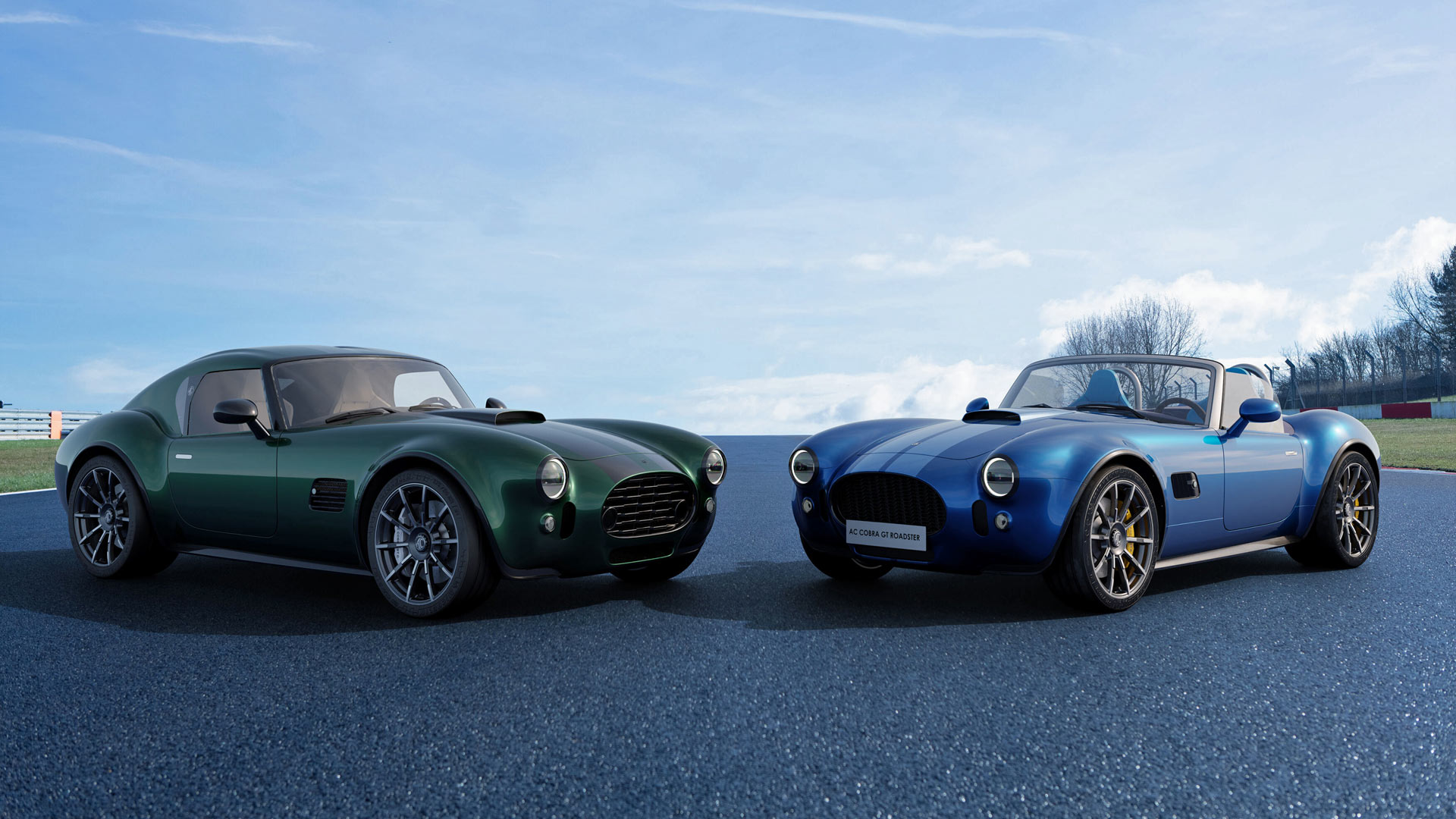 Two vintage-style sports cars, one green and one blue, parked on a sunny race track. Leafless trees and clear skies surround.