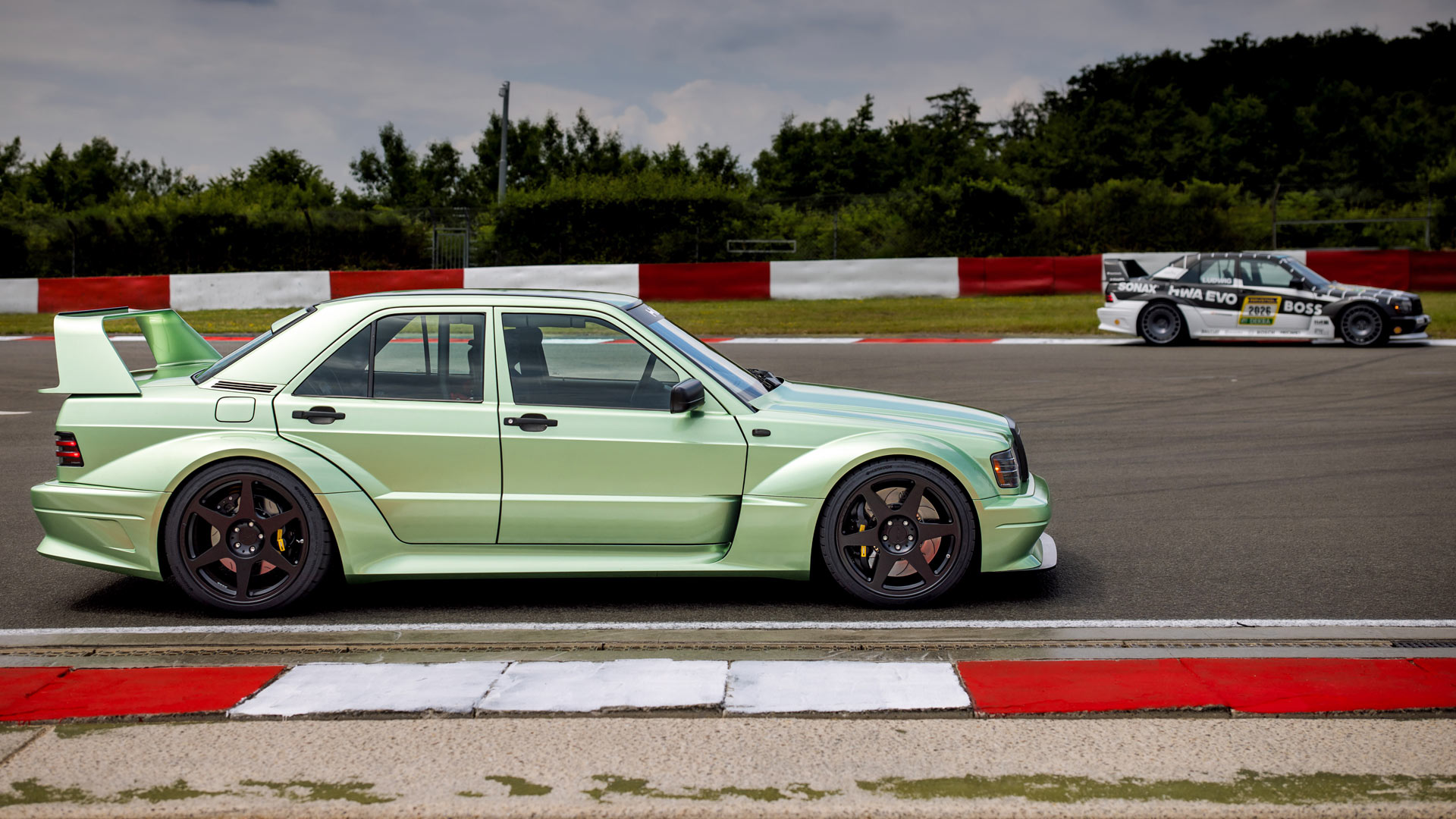 Two HWA Evo cars on a race track with red and white barriers, surrounded by trees under a cloudy sky.
