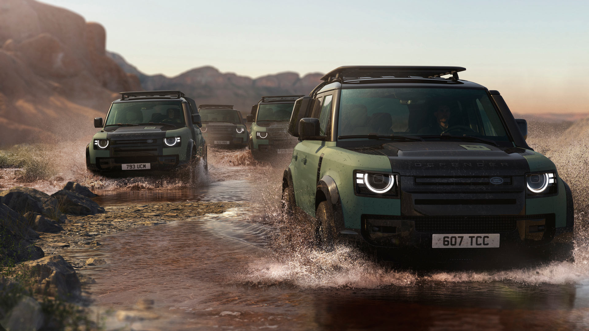 Four Land Rover Defenders drive through a shallow, rocky stream in a desert landscape, with rugged mountains in the background.