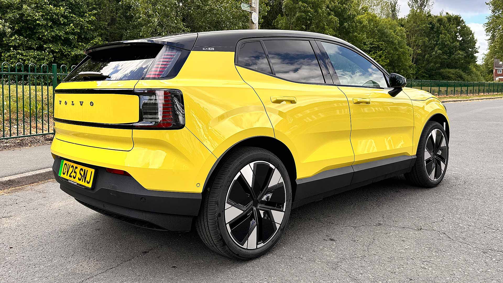 A yellow Volvo EX30 SUV is parked on a road beside greenery and a fence, under a partly cloudy sky.