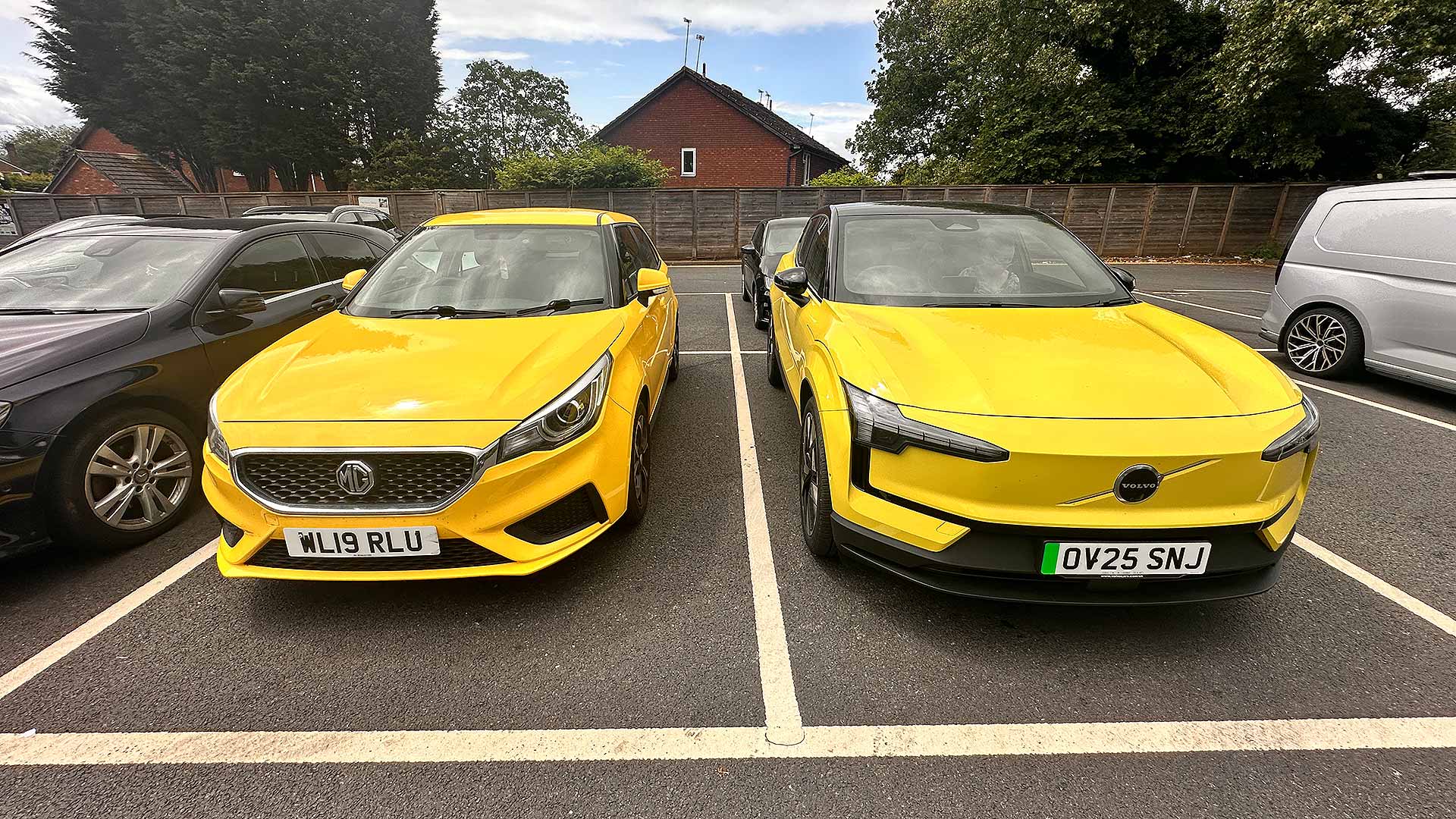 Two yellow cars parked side by side, one is MG, the other Volvo. Background shows a wooden fence and brick house.
