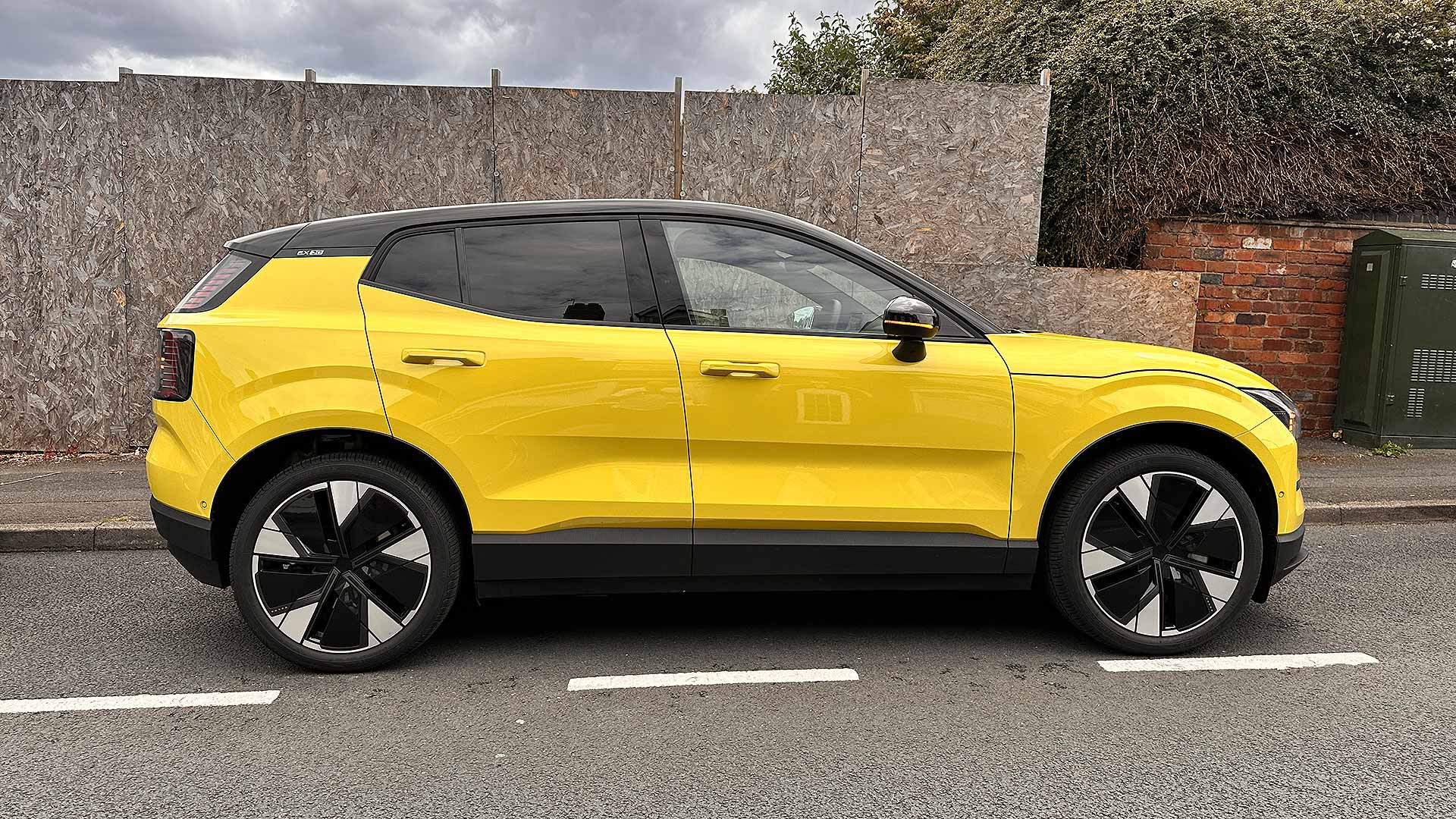A bright yellow SUV parked on a residential street, with a wooden fence and brick wall in the background on an overcast day.